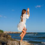 A woman in a yoga pose on a rock by the ocean, enjoying a sunny day.